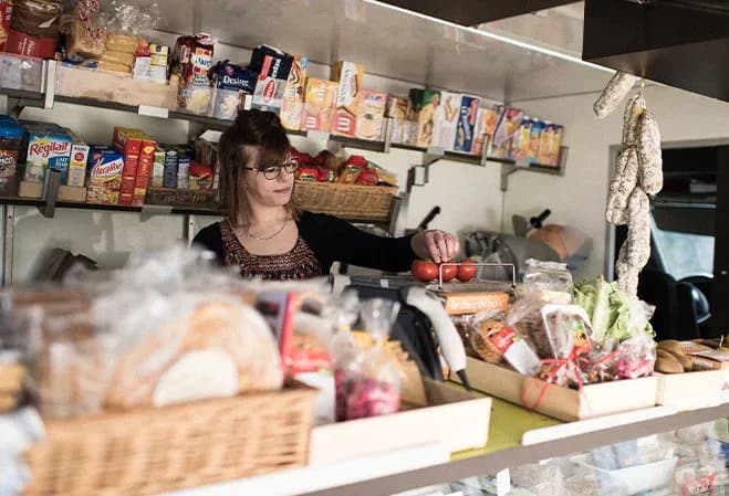 femme dans une épicerie pesant des tomates entourée de produits alimentaires variés