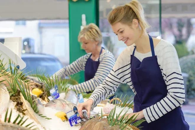 jeune femme souriante prépare des poissons sur un étal de marché avec une collègue en arrière-plan