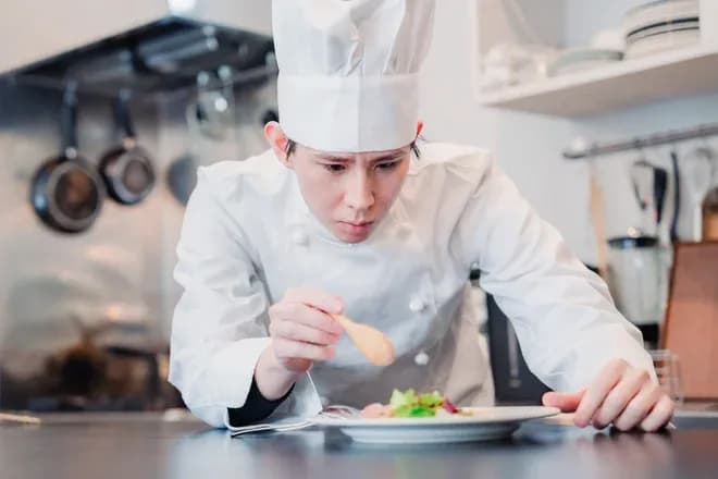 chef en uniforme examinant un plat avec une cuillère dans une cuisine moderne
