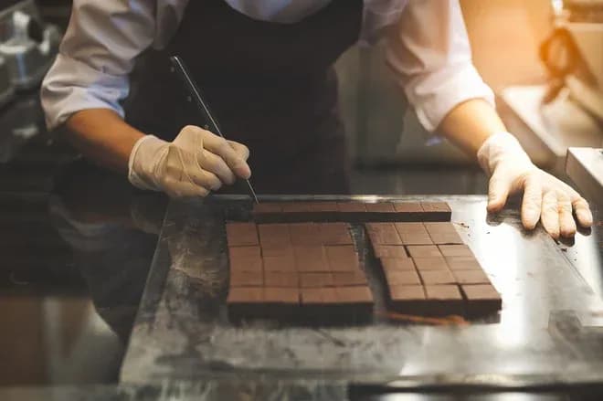 artisan chocolatier ganté découpant des carrés de chocolat sur plan de travail