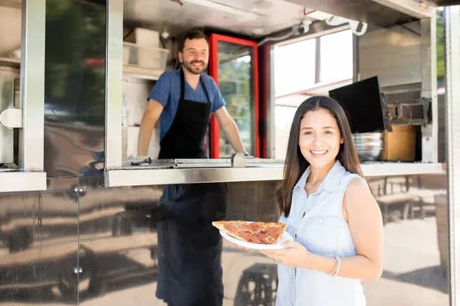 Femme souriante tenant une part de pizza devant un camion de restauration rapide avec un serveur.
