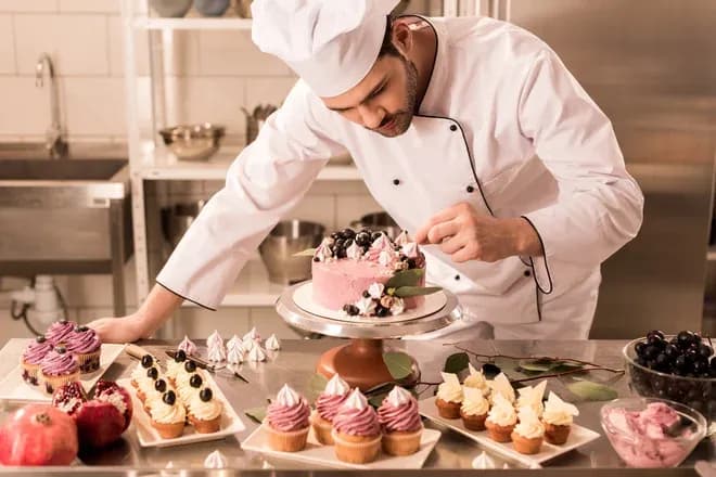 chef pâtissier décorant un gâteau rose entouré de cupcakes et meringues en cuisine