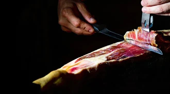 main-d'une personne coupant du jambon avec un couteau sur une planche en bois