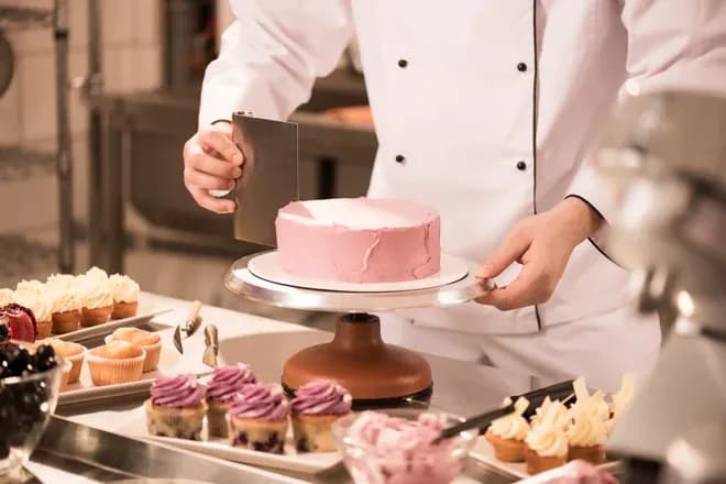 chef pâtissier lissant un glaçage rose sur un gâteau entouré de cupcakes