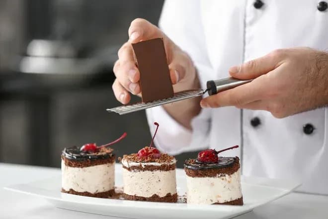 mains d'un pâtissier râpant du chocolat sur trois entremets aux cerises