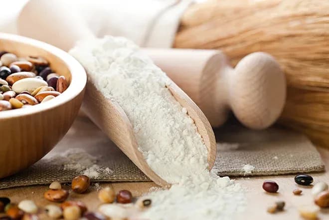 pelle en bois renversant de la farine près d'un bol de haricots secs et d'un rouleau