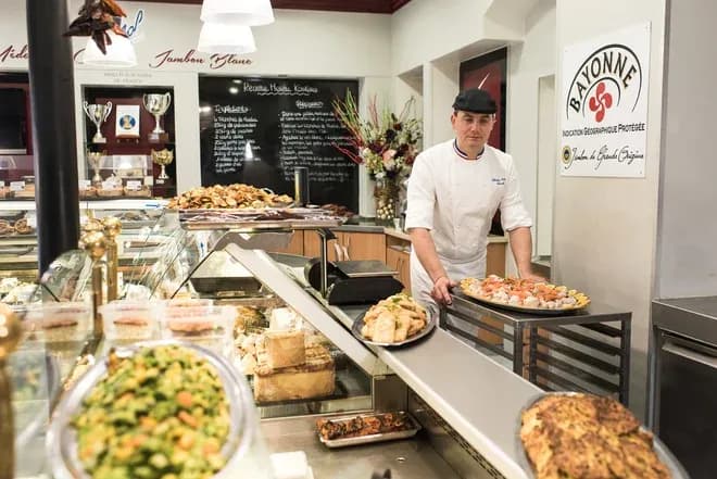 Traiteur en blouse et béret présentant plateaux de mets derrière un comptoir