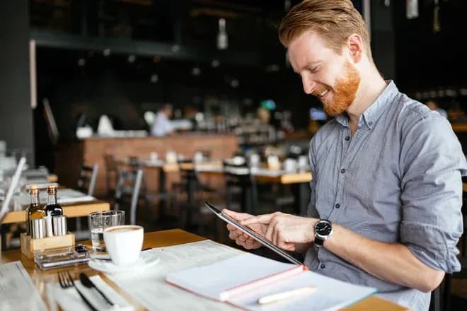 Homme souriant utilisant une tablette dans un restaurant avec un café et un carnet devant lui