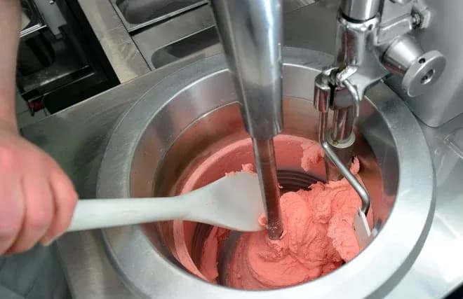 main avec spatule raclant une glace rose dans une machine à glace professionnelle