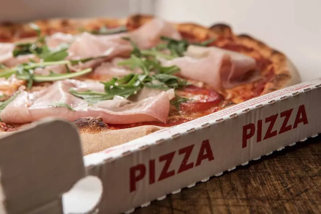 pizza garnie de jambon et roquette dans une boîte avec le mot pizza en rouge