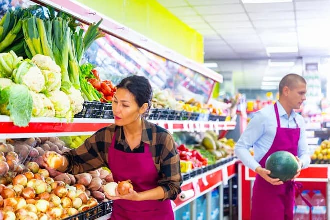 femme choisissant des légumes dans un magasin avec un homme portant une pastèque en arrière-plan