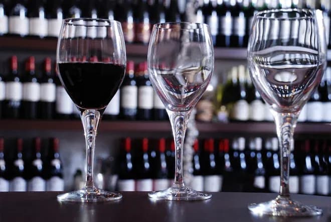 trois verres à vin dont un rempli de vin rouge devant étagères de bouteilles