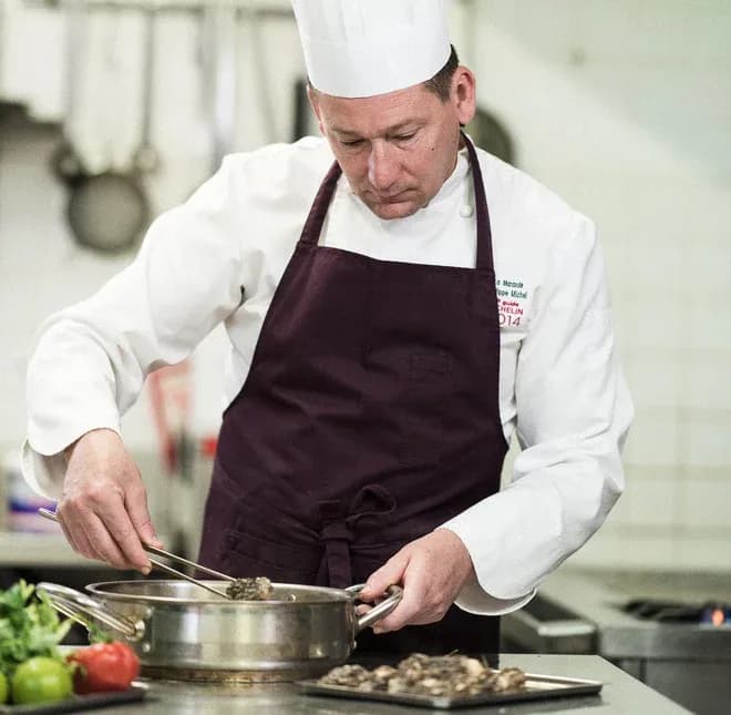 chef en toque et tablier dépose des ingrédients dans une poêle en cuisine professionnelle