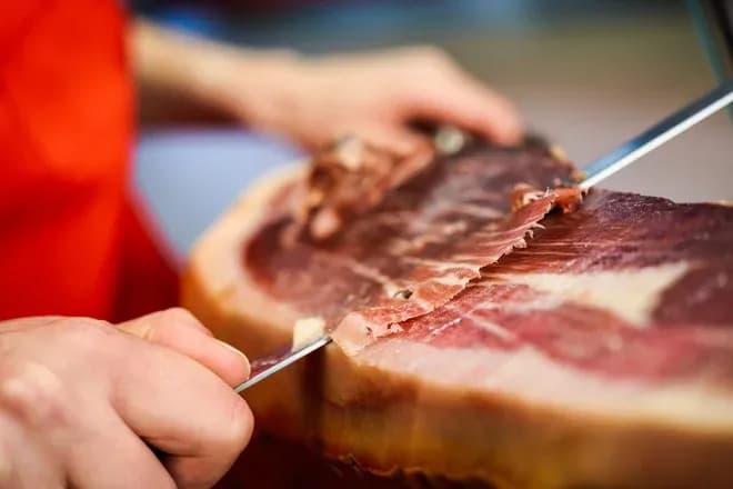 main-d'une personne coupant du jambon avec un couteau sur une planche à découper