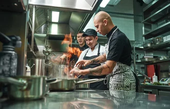 trois cuisiniers en cuisine professionnelle préparant un plat flambé