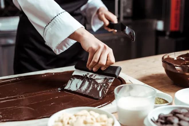 artisan chocolatier étale du chocolat fondu sur une surface en bois avec divers ingrédients autour