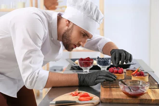 pâtissier coiffé d'une toque et gants noirs disposant des framboises sur tartelettes