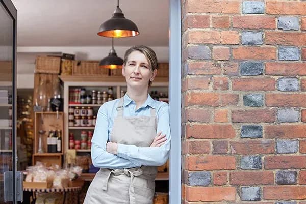 femme souriante en tablier devant une épicerie avec des étagères remplies de produits