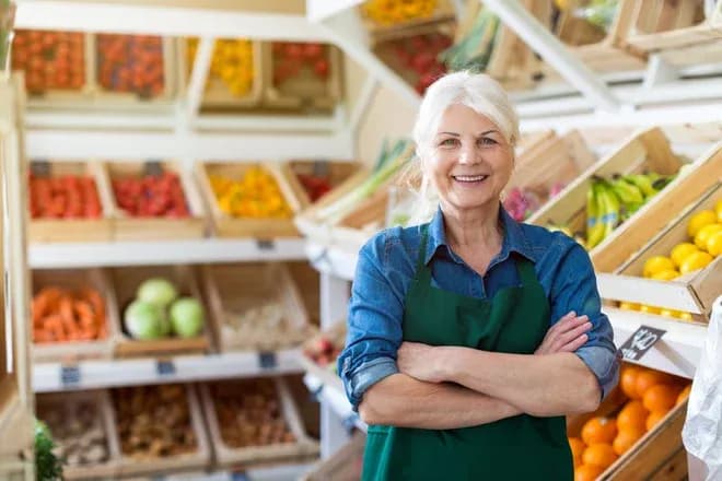 Femme souriante en tablier devant des étals de fruits et légumes dans un magasin.