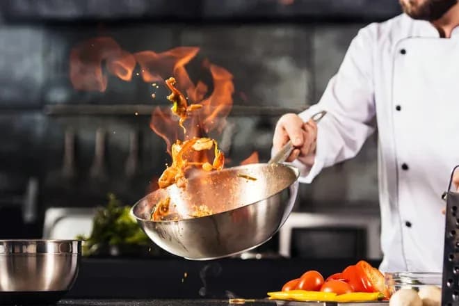 chef en veste blanche faisant flamber des légumes dans une poêle