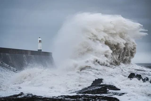 vague massive s'écrasant contre un mur avec un phare en arrière-plan sous un ciel nuageux