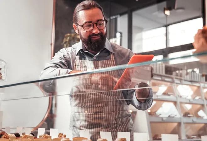 homme souriant en tablier utilisant une tablette devant une vitrine de pâtisseries