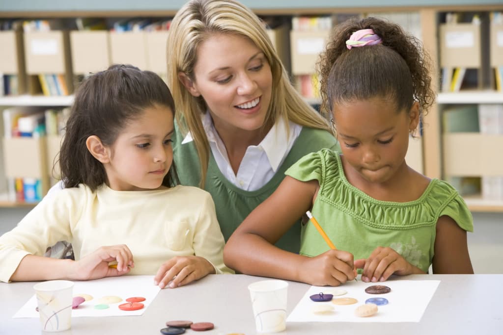 enseignante accompagnant deux enfants en train de dessiner à une table avec des fournitures d'art