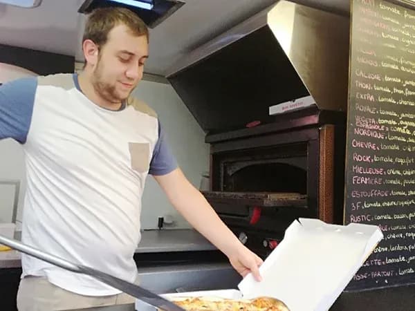 un homme souriant sort une pizza d'une boîte dans un restaurant avec un four à pizza en arrière-plan