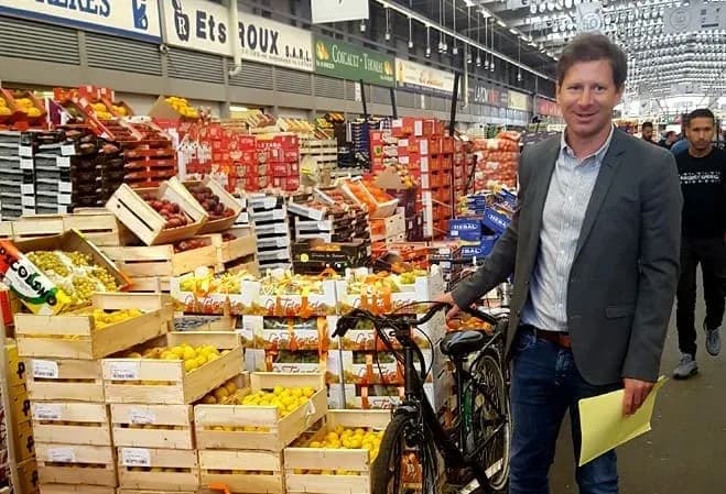 Homme souriant avec un vélo au milieu d'un marché coloré de fruits et légumes