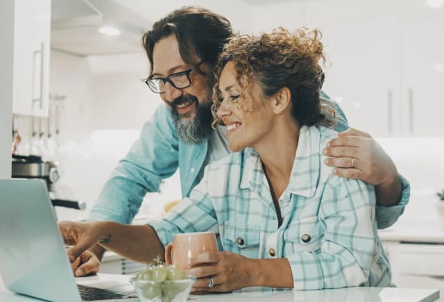couple souriant devant un ordinateur portable dans une cuisine moderne avec une tasse à la main