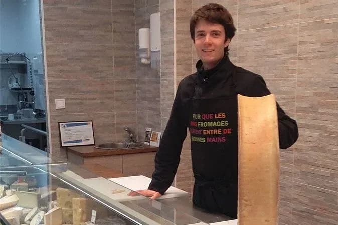 Homme souriant dans une fromagerie tenant un grand morceau de fromage devant un comptoir