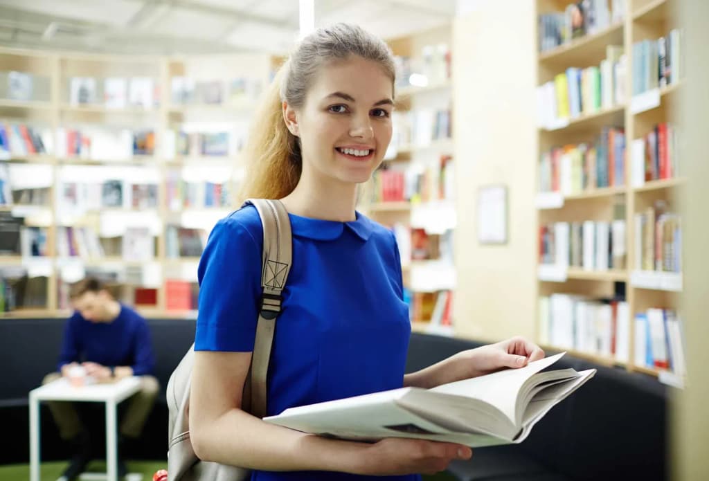étudiante souriante tenant un livre dans une bibliothèque avec des étagères remplies de livres