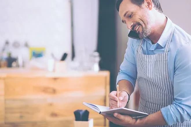 homme souriant au téléphone prenant des notes dans un carnet de cuisine dans un environnement chaleureux