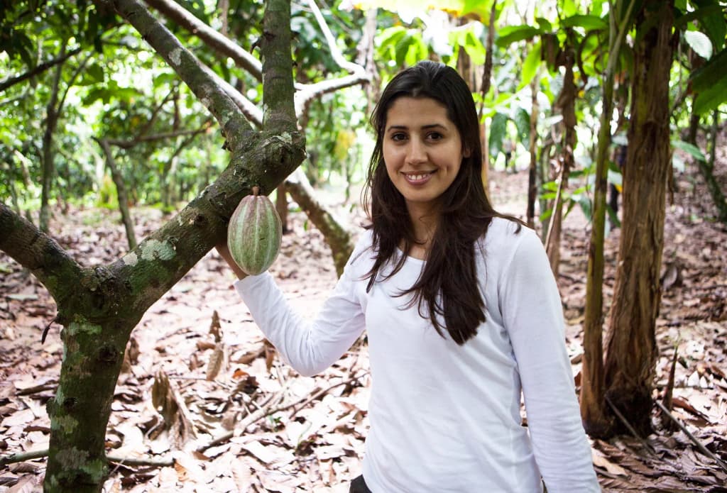 femme souriante dans une plantation de cacao tenant une cabosse de cacao entre les arbres