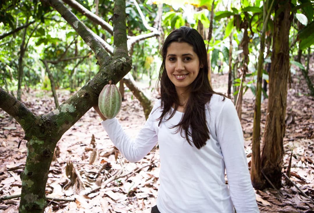 femme souriante dans une plantation de cacao tenant une cabosse de cacao entre les arbres