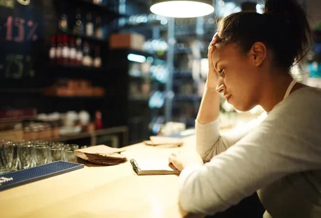 femme pensive dans un café avec une main sur le front et un carnet devant elle