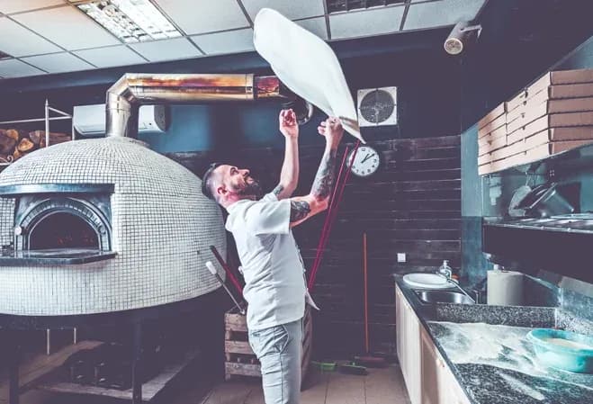 Un pizzaiolo étire une pâte à pizza dans une cuisine avec un four à bois en arrière-plan