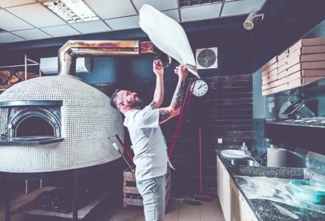 Un pizzaiolo étire une pâte à pizza dans une cuisine avec un four à bois en arrière-plan