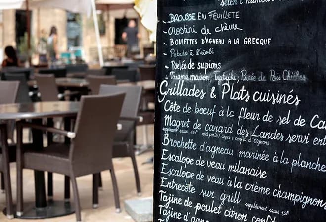 menu écrit à la craie sur un tableau noir devant une terrasse de restaurant avec des tables en extérieur