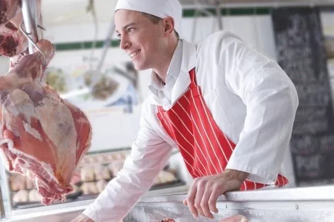 boucher souriant travaillant derrière un comptoir avec des morceaux de viande suspendus