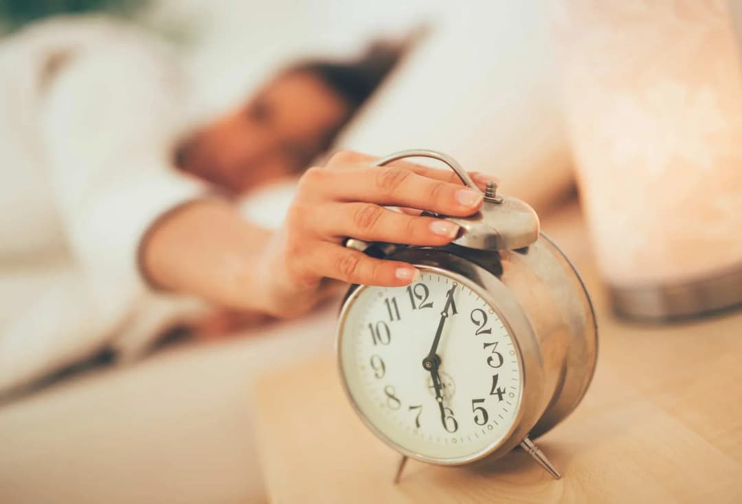 main d'une personne éteignant un réveil sur une table près d'un lit avec une lampe douce