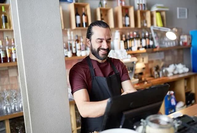 homme souriant utilisant un logiciel de caisse dans un café avec des bouteilles en arrière-plan