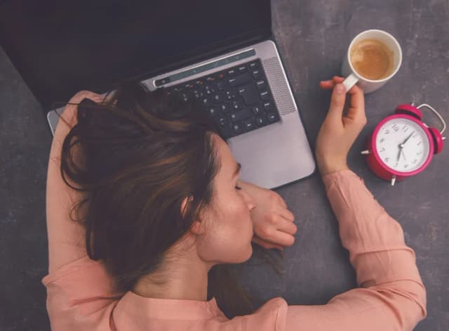 femme fatiguée s'appuyant sur un ordinateur portable avec une tasse et un réveil à côté