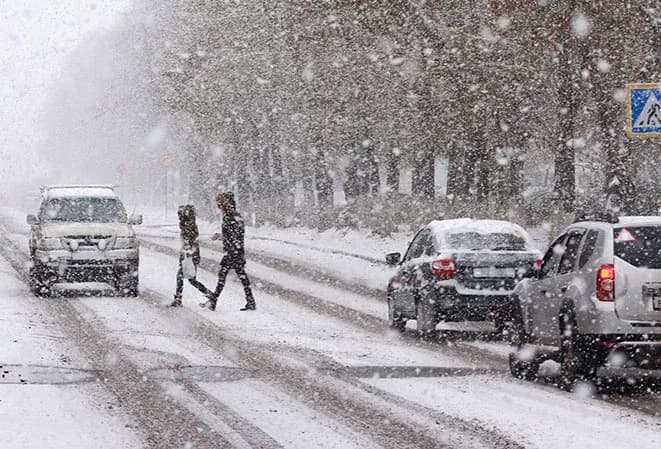 Deux personnes traversent une route enneigée sous une forte chute de neige avec des voitures garées.