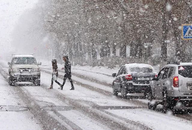 Deux personnes traversent une route enneigée sous une forte chute de neige avec des voitures garées.