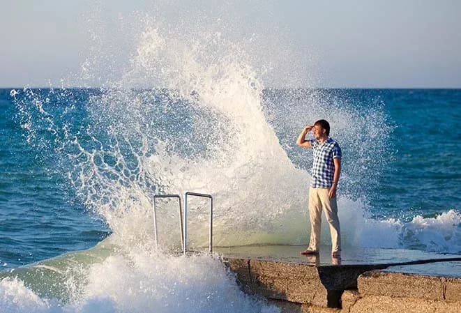 homme sur un quai face à une grande vague qui se brise contre le rocher