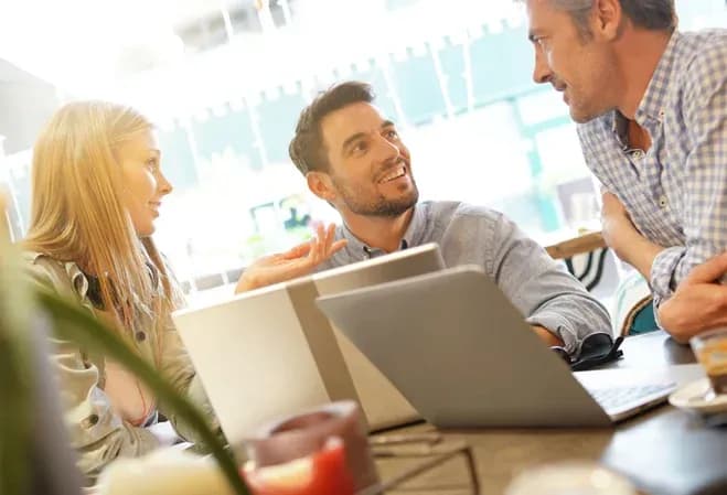 Trois personnes discutent autour d'une table avec des ordinateurs portables et des boissons.