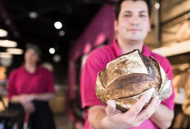 un boulanger présente un pain dans une boulangerie avec un collègue en arrière-plan