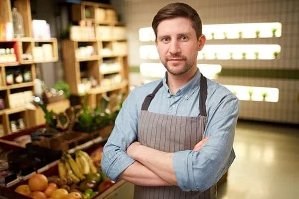 Homme souriant en tablier devant des étagères de fruits et légumes dans un magasin.
