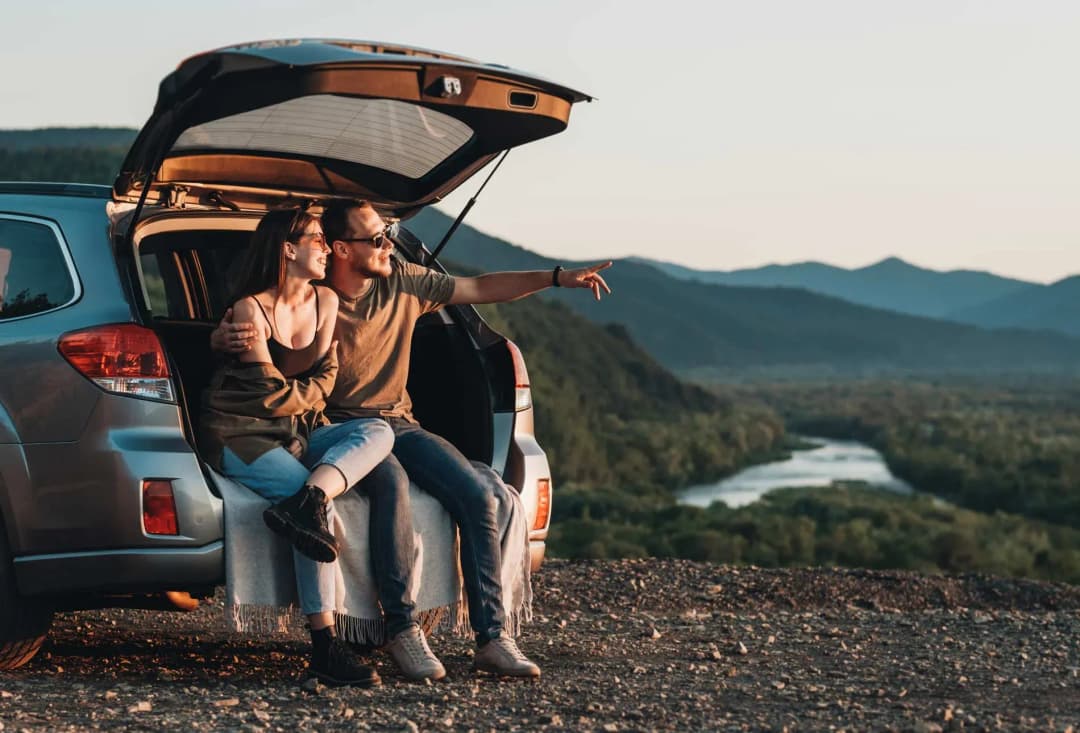 couple assis sur le bord d'une voiture avec vue sur une vallée et des montagnes au coucher du soleil